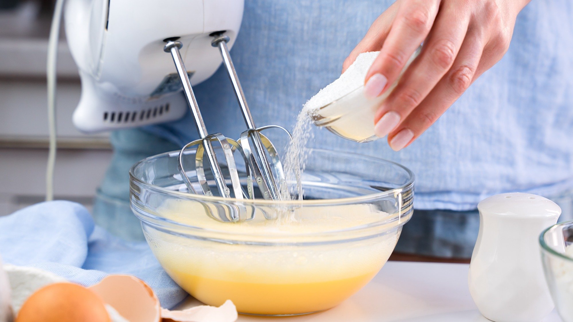 Рука насыпает сахар в миску с яйцами, которые перемешиваются ручным миксером